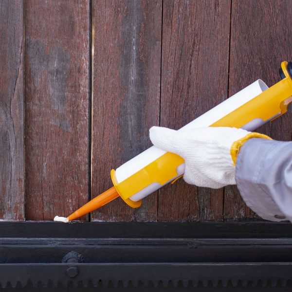 Close-up of a hand in a white glove using a caulk gun to seal a gap near a wooden door frame, illustrating pest exclusion.
