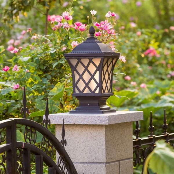 A decorative outdoor lamp post in a garden setting, surrounded by pink flowering shrubs and an ornate iron fence.