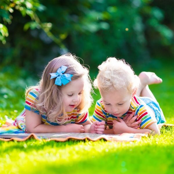 A young girl and boy, both with brightly colored striped shirts, lying on a blanket on a sunny, green lawn, looking closely at a book or map.