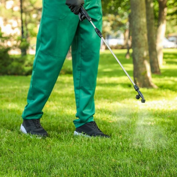 Close-up of a technician's legs and shoes in green uniform pants, applying liquid treatment to a green lawn with a wand sprayer.