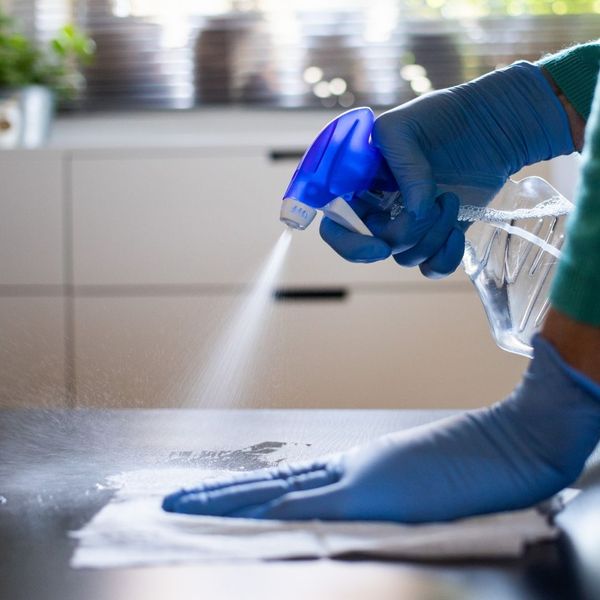 Close-up of hands in blue gloves spraying disinfectant onto a dark kitchen counter surface to sanitize the area.