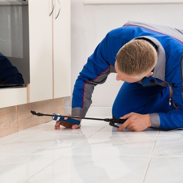 A pest control technician in a blue uniform kneeling on a white tiled floor, applying targeted treatment with a thin wand sprayer along the base of a kitchen cabinet.