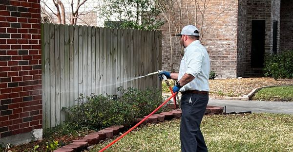 A pest control technician wearing a hat and light shirt spraying a red hose along a wooden fence and brick wall in a residential yard. A pest control technician wearing a hat and light shirt spraying a red hose along a wooden fence and brick wall in a residential yard.