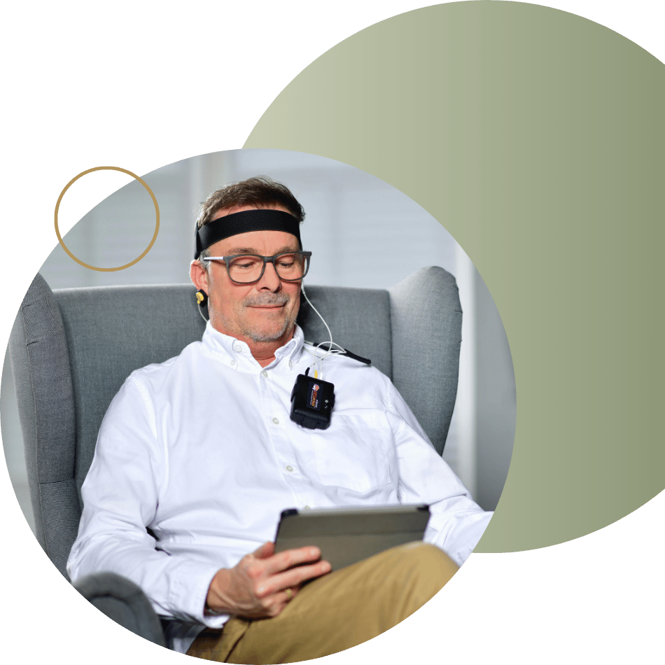 Man in a chair wearing a biofeedback or neurofeedback device on his head, looking at a tablet, representing technology-assisted anxiety therapy.