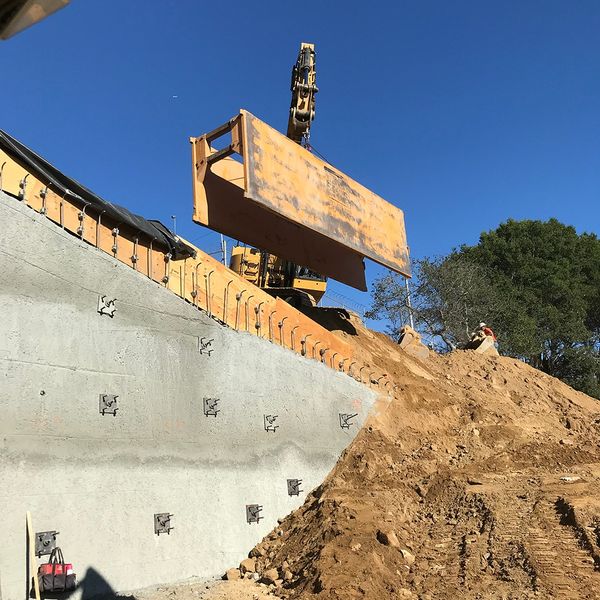 Excavator lifting a large trench shield into place near a newly reinforced concrete retaining wall.