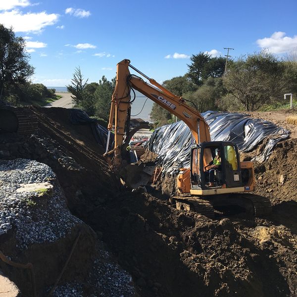 Orange excavator digging a deep, precise utility trench alongside a coastal road.
