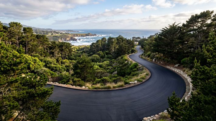 A beautiful, finished asphalt driveway in Monterey surrounded by greenery and coastal trees under a clear sky.