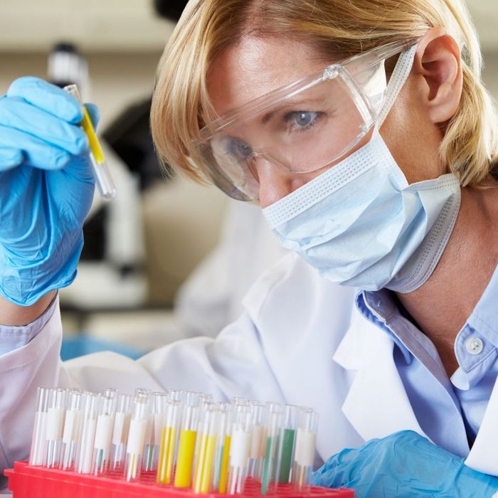 A female scientist in a laboratory wearing a face mask, safety goggles, and blue gloves, carefully examining a test tube containing a yellow liquid.