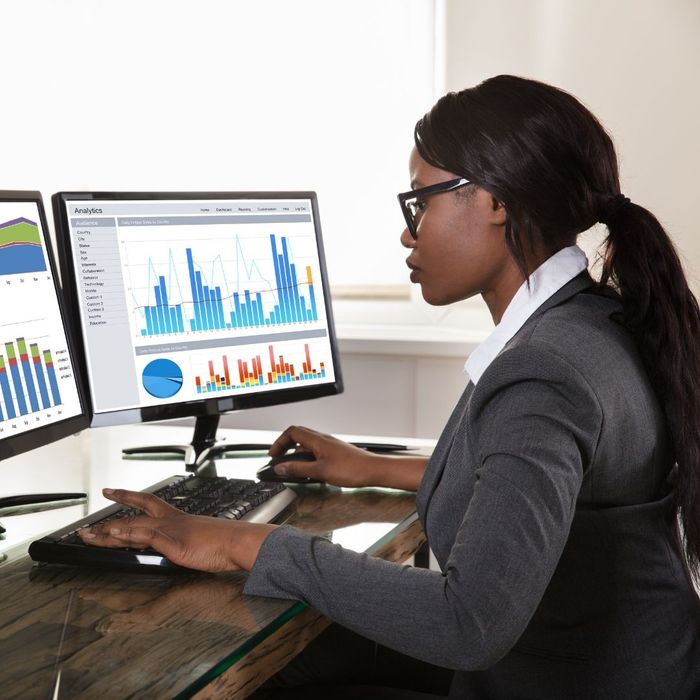 A professional Black woman with glasses working at a desk, focused on dual computer monitors displaying various colorful data analytics and business charts.