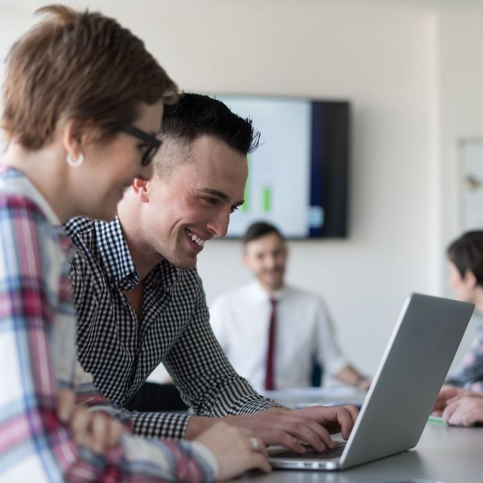 A man and woman sitting together at a desk, smiling and laughing while looking at a laptop screen during a collaborative work session.