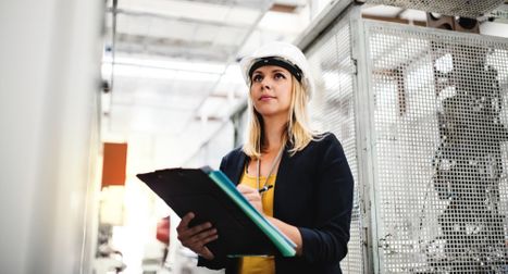 A blonde female engineer wearing a white hard hat and a blazer, holding a clipboard and looking upward thoughtfully inside a modern industrial or technical facility.