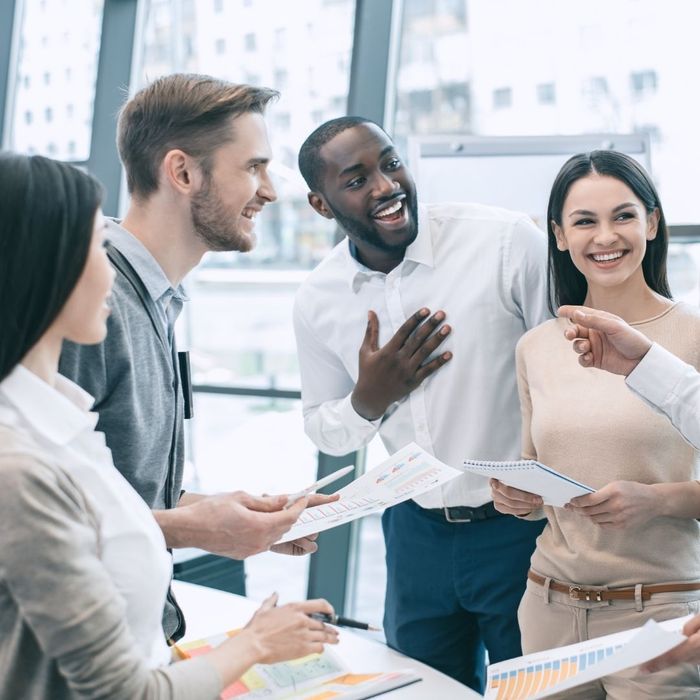 A diverse group of four professionals standing in a bright office, laughing and talking while holding notebooks and reviewing data charts.