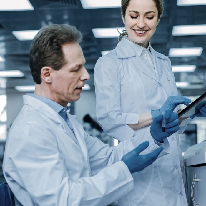 A female laboratory professional in a white coat smiling while using a digital tablet, standing next to a male colleague who is holding a small sample vial.