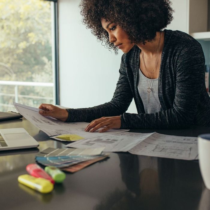 A woman with curly hair leaning over a dark table, meticulously reviewing architectural blueprints and color swatches in a brightly lit workspace.
