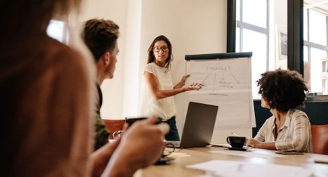 A woman wearing glasses stands at the head of a conference table, pointing to a strategic "Strategy" flowchart on a flip chart during a team meeting.