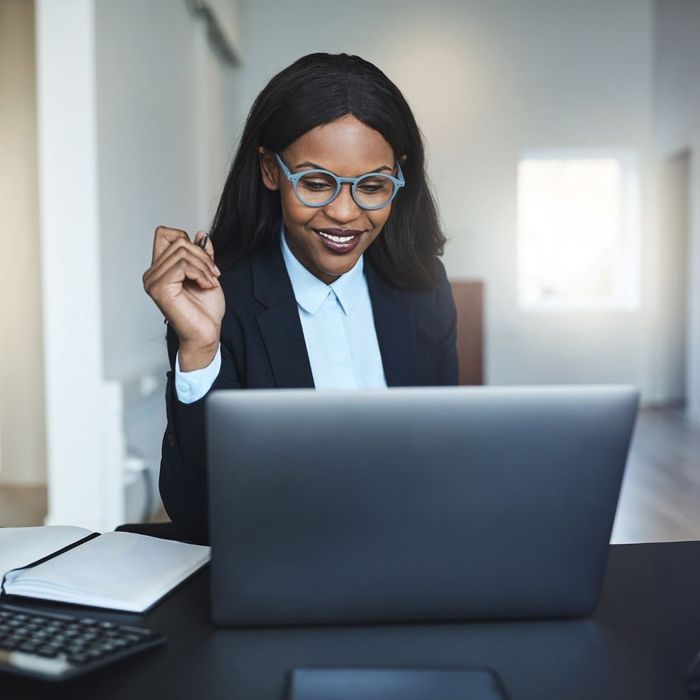 A Black businesswoman with stylish blue glasses smiling while working on a laptop at her desk in a bright, modern office setting.