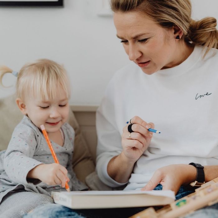 A mother in a white sweatshirt sits closely with her young toddler, both focused on coloring together in a book with colored pencils.