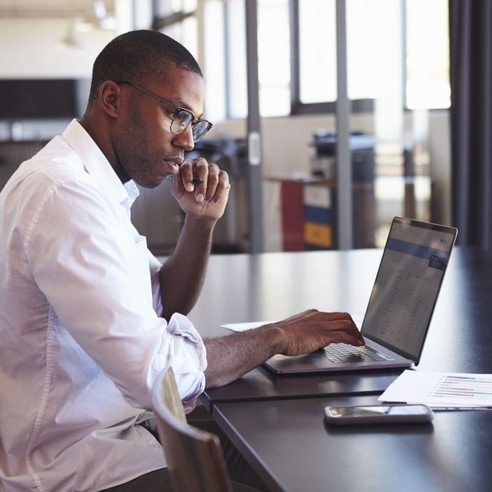 A professional man with glasses and a white shirt sitting at a long dark table, deeply focused while typing on his laptop in a modern office.