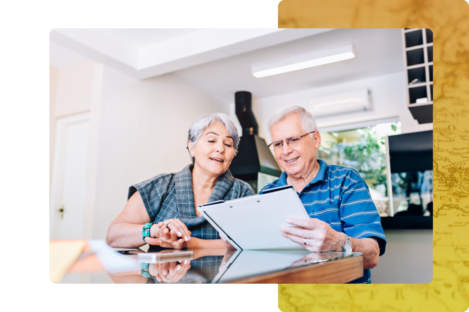 An elderly couple sitting at a dining table, looking closely at a tablet or folder together with a focused and positive expression.