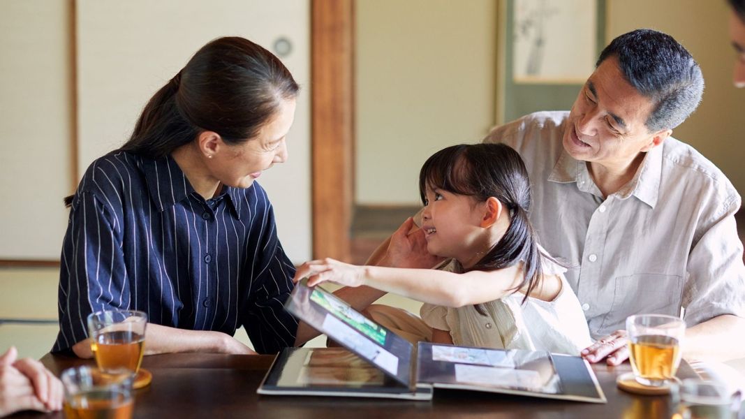An intimate scene of a multi-generational family—a grandmother, grandfather, and young granddaughter—smiling together while looking through a photo album at a table. An intimate scene of a multi-generational family—a grandmother, grandfather, and young granddaughter—smiling together while looking through a photo album at a table.