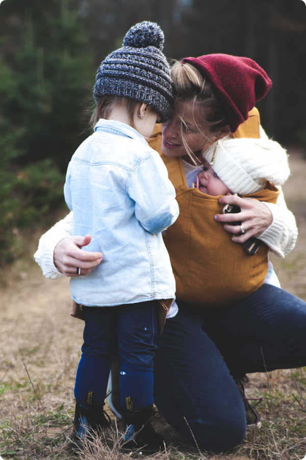 a mother, wearing a red beanie, kneels to embrace her young child while carrying an infant in a mustard-colored baby carrier.