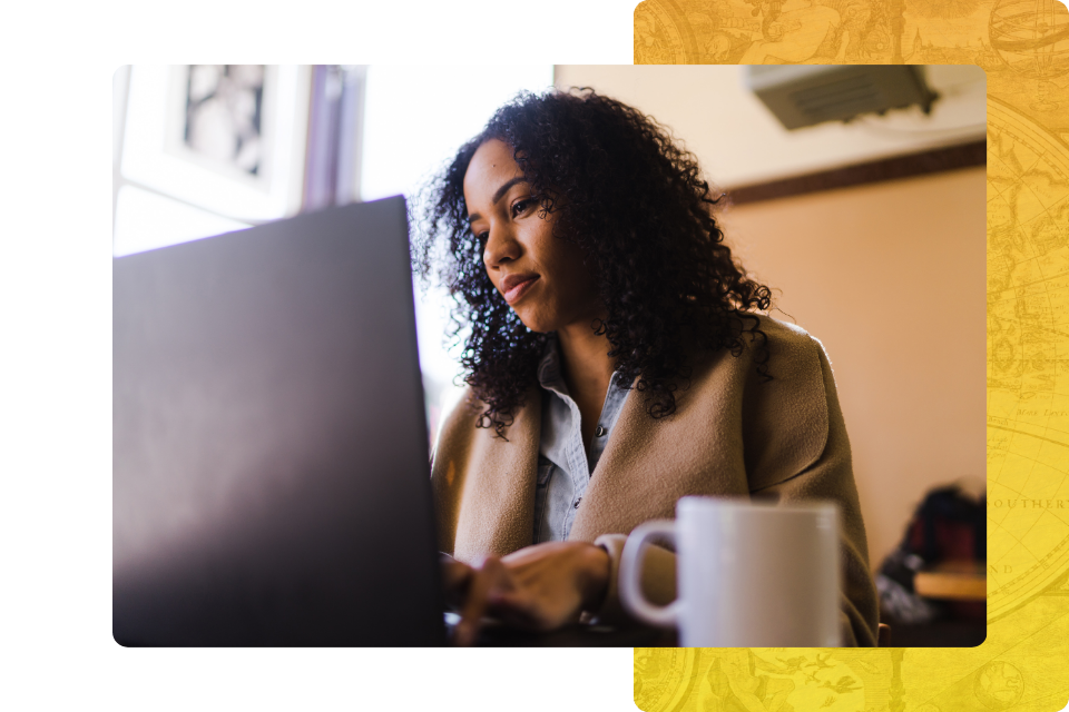 A young woman with curly hair looking focused while working on a laptop at a table, with a warm cup of coffee in the foreground.