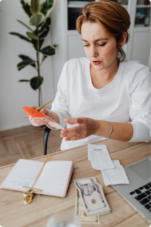 A woman at a desk looking concerned or focused while reviewing receipts and financial documents next to a stack of cash and an open planner.