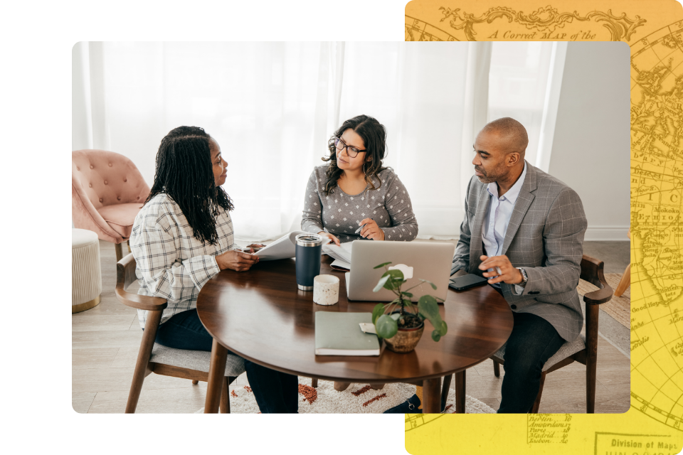 two women and one man sitting around a circular wooden table in a bright, modern office, engaged in a collaborative discussion over open files.