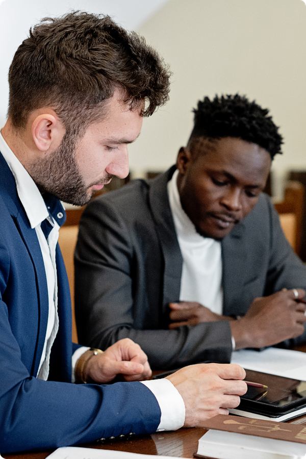 Two professional men sitting at a wooden table, focused intensely on a tablet and documents during a serious business consultation.