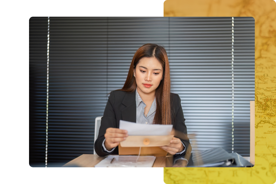 A professional woman in a black blazer sitting at a desk, carefully reviewing a document she has just pulled from a large manila envelope.