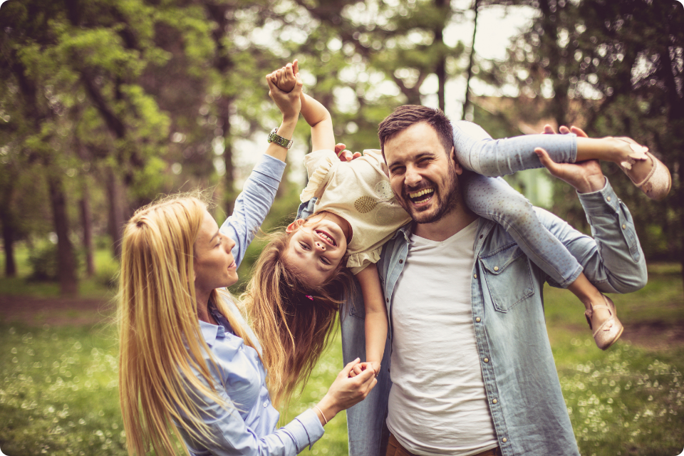 a happy father holding his young daughter upside down on his shoulders while the mother laughs and holds the child's hand in a sunlit park.