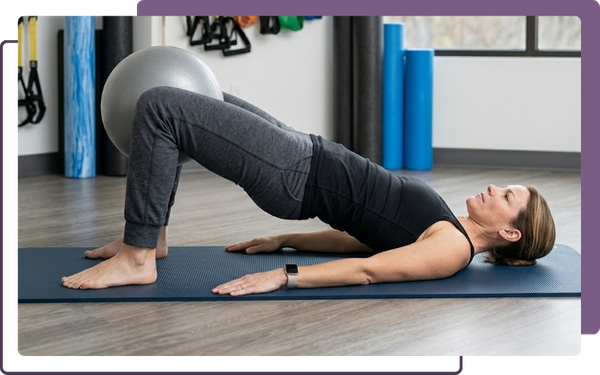 A 1:1 professional photograph capturing the active female patient performing a controlled glute bridge stabilization exercise while holding a gray exercise ball between her knees on a mat in a bright, modern clinical gym setting.