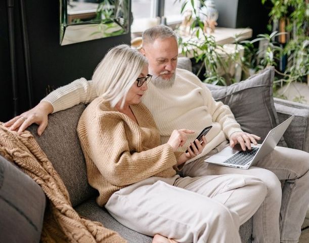 A senior couple sitting comfortably on a sofa together, looking at a laptop and smartphone, representing retirement planning
