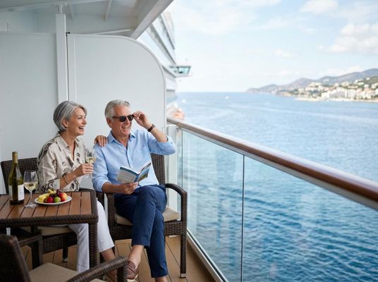 A happy older couple enjoys drinks and a book on a cruise ship balcony with a scenic ocean and distant coastline view. Happy Couple on Cruise Ship Balcony