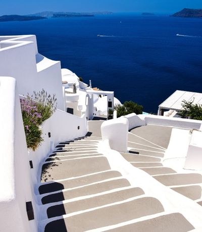 A white staircase with grey steps winds down past whitewashed buildings towards the deep blue sea, with islands visible in the distance under a clear sky.