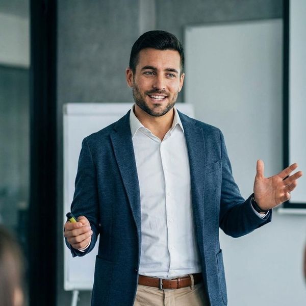 A confident man in business casual attire speaking and gesturing with a smile during a presentation.