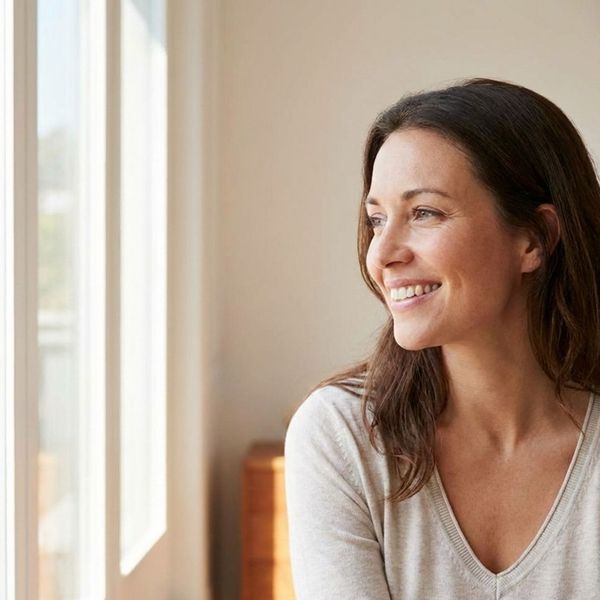 A woman with a relaxed, natural smile looking out a large window in a sunlit room.