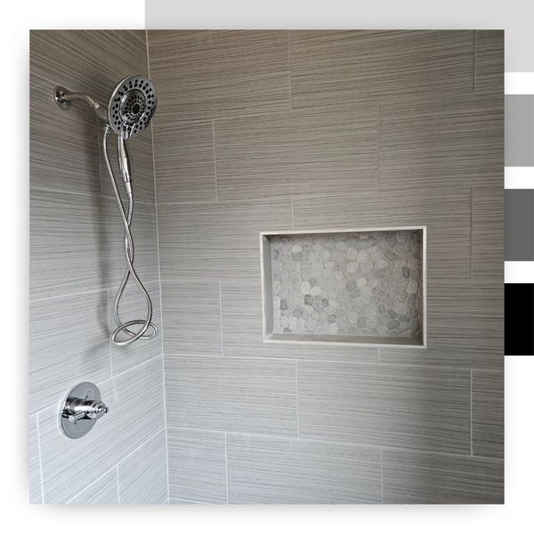 A close-up of a modern shower wall featuring long, horizontal gray textured tiles. A chrome handheld shower head fixture is on the left, and a large, recessed shower niche is built into the wall with a contrasting light gray pebble mosaic tile backer.