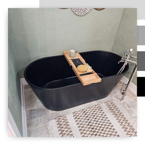 A luxurious, contemporary black freestanding soaking tub set against a textured sage green wall, with a light-colored woven rug on the gray tiled floor.