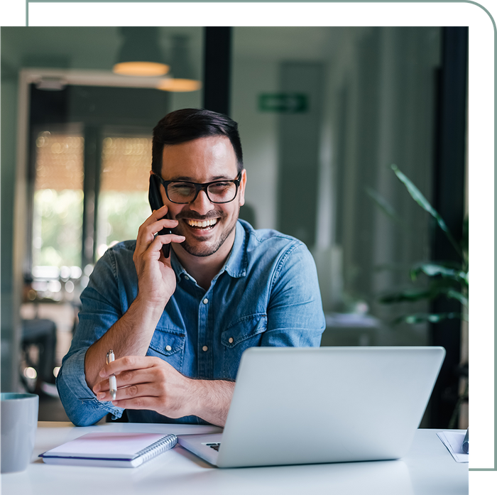 man sitting with pen and paper while happily talking on the phone and using a laptop