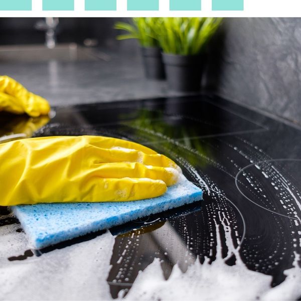 Close-up of a person in yellow rubber gloves using a blue sponge to scrub a black glass stovetop with soapy suds.