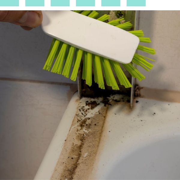 Extreme close-up of a white scrubbing brush cleaning deep grime and dirt from a bathroom tile corner.