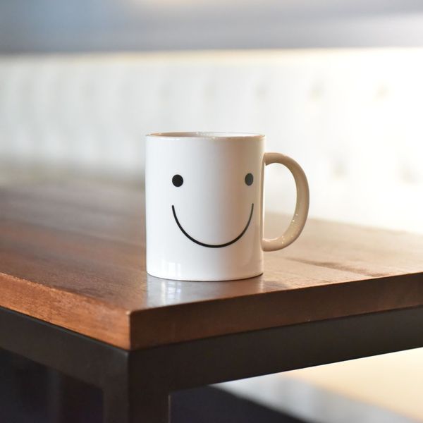 A white coffee mug with a black smiley face design sits on the corner of a wooden table Best Amenities for Remote Workers 1.jpg