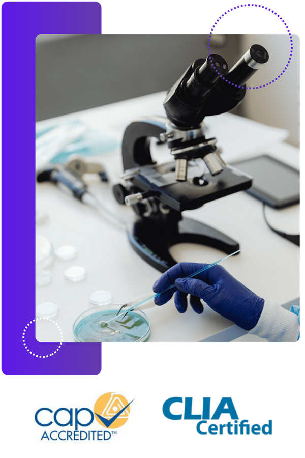 A gloved hand holds a tool over a petri dish with green liquid, with a microscope and other lab equipment on the table.