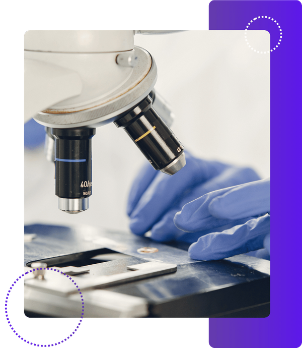 A close-up shot of a microscope with two objective lenses visible. A gloved hand is adjusting a slide on the microscope stage.