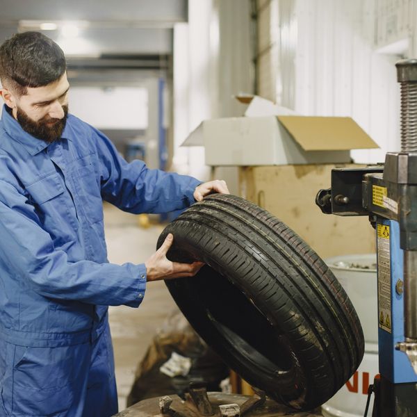 A male mechanic in blue coveralls carefully inspects the tread of a tire inside an auto repair shop.