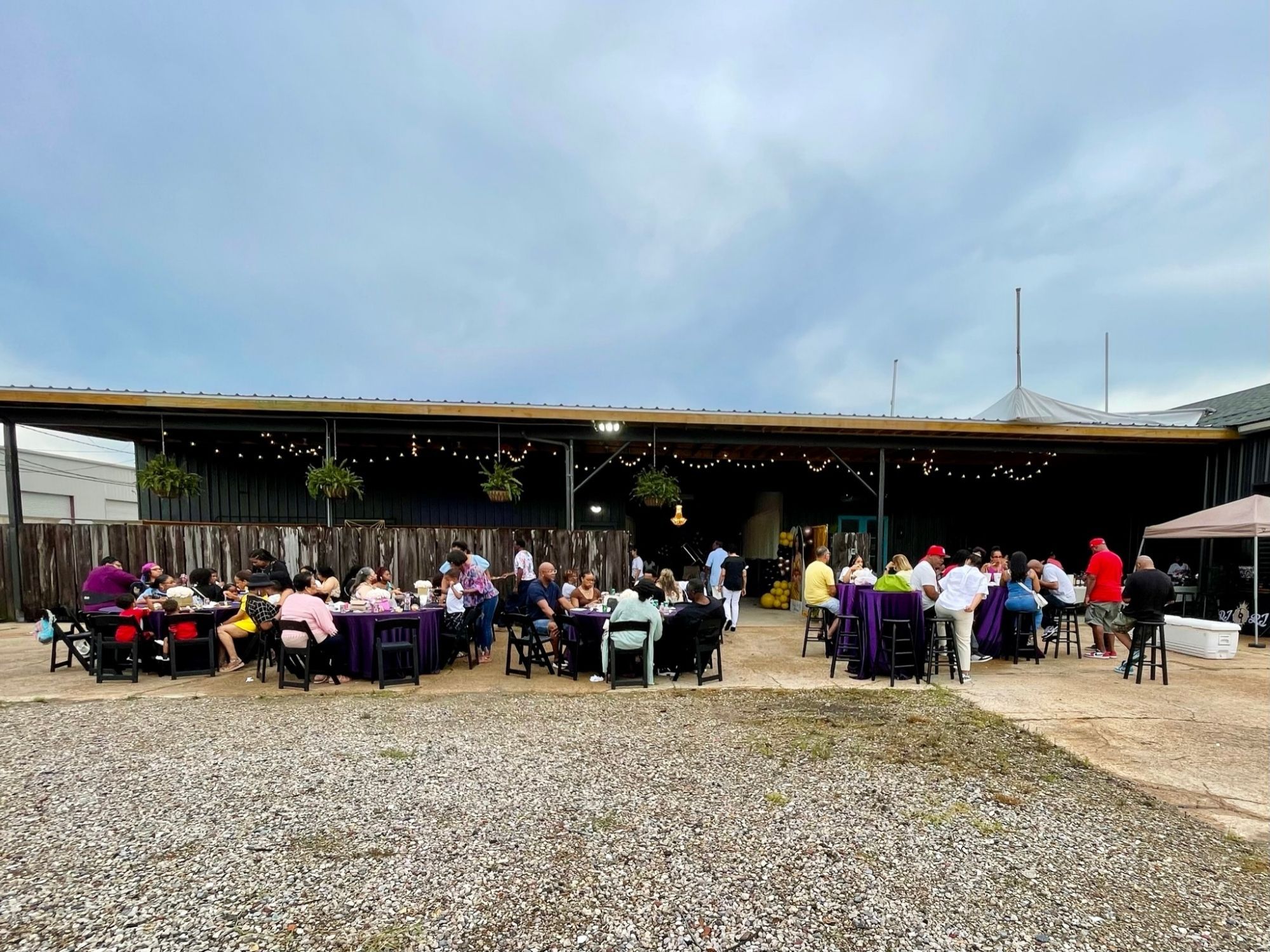 Wide view of NOLA Station's casual venue with guests seated at purple high-top tables in the outdoor gathering space under a long covered patio with string lights