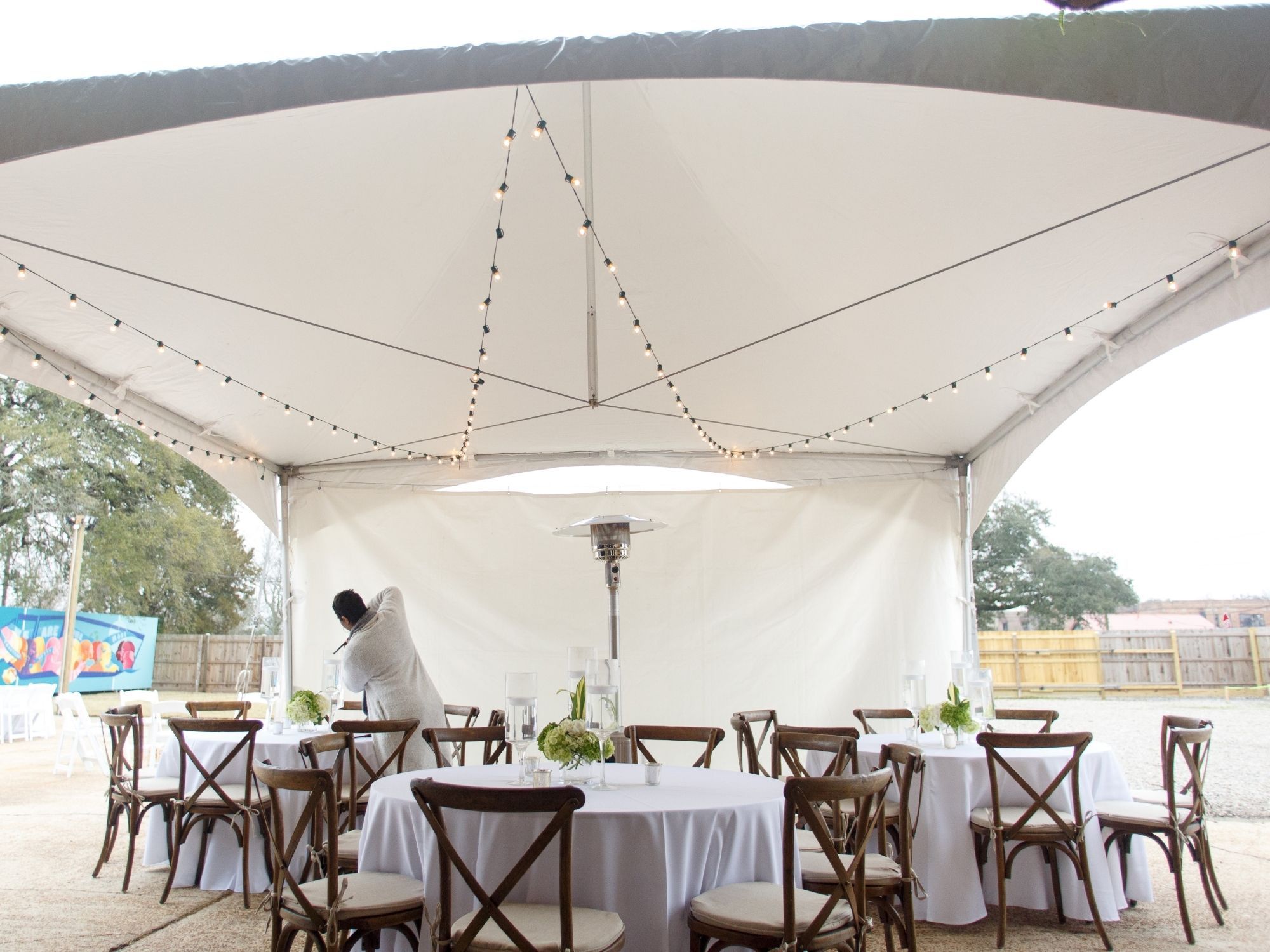 White linen round tables and wooden chiavari chairs set up beneath a large white event tent with string lighting White linen round tables and wooden chiavari chairs set up beneath a large white event tent with string lighting