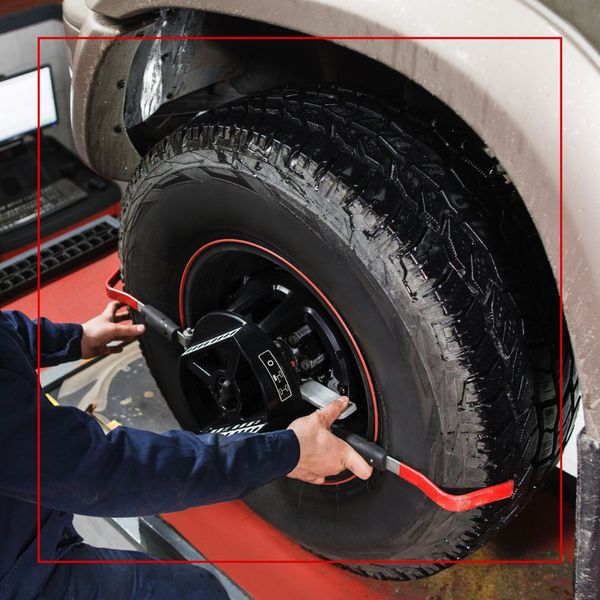 A mechanic in blue overalls is adjusting a wheel alignment clamp attached to a large, textured tire on a vehicle. A mechanic in blue overalls is adjusting a wheel alignment clamp attached to a large, textured tire on a vehicle.