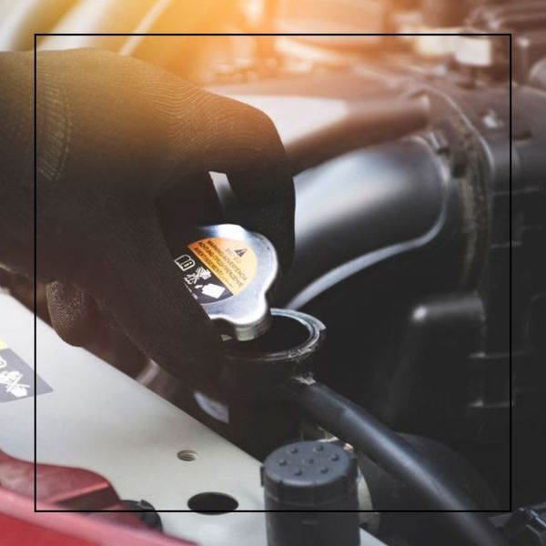 A gloved hand unscrews the cap of a car's radiator, with engine components and a red car body visible in the background, suggesting maintenance or inspection.